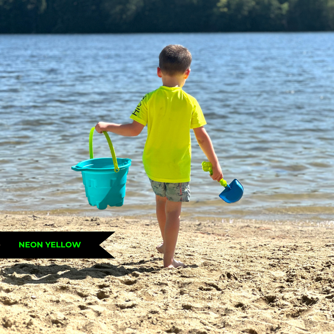 Photo of a boy walking away from the camera toward the edge of the water on a beach. Carrying a bucket and shovel. He is wearing a neon yellow rash guard short-sleeve and a pair of pale blue swim trunks.